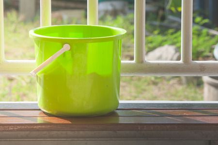 Green plastic water bucket or pail put on wooden long chair with white steel fence in the background. (Selective focus)の写真素材