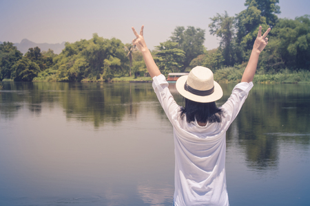 Asian woman wear hat and white shirt with standing on wooden bridge. She looking forward to river with raising hands up and she has feeling relax and happiness.の写真素材