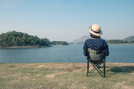 Vacation and Travel Concept : Asian woman wear hat and sitting relax on portable chair nearly lake at National Park.の写真素材