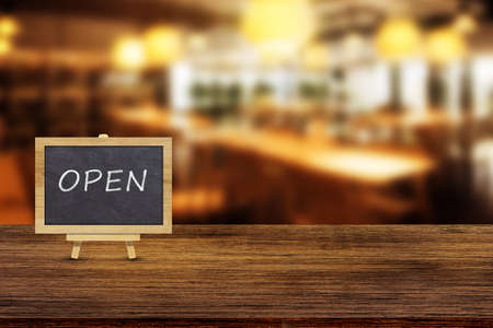 Open signs text on wooden board with blurry image of restaurant or coffee shop in background.
