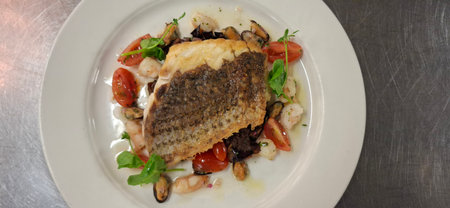 Elevated seafood dish showcasing pan-seared fish atop a medley of shrimp, mussels, cherry tomatoes, and garnished with fresh greens, all plated beautifully for gourmet dining or culinary design purposes.の写真素材