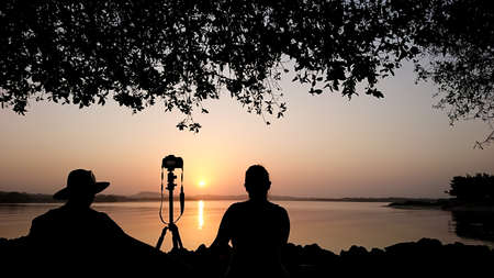 Young couple sitting on a rock near the sea making photos of the dawn, silhouette of a photographer...