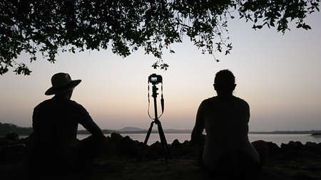 Young couple sitting on a rock near the sea making photos of the dawn, silhouette of a photographer...