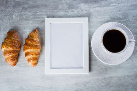 Blank white frame with copy space, cup of coffee and croissants on a grey table background . Mockup with copy space, top viewの写真素材