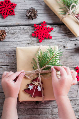 Girl's hands wrap christmas holiday handmade present in craft paper with red tree. Christmas gift on wooden rustic table background. Flat lay, top viewの写真素材