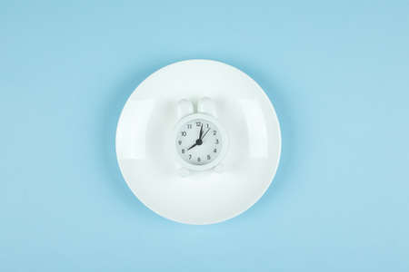 Intermittent fasting, ketogenic, diet concept. Empty plate with white alarm clock on blue background. Flat lay, top viewの写真素材
