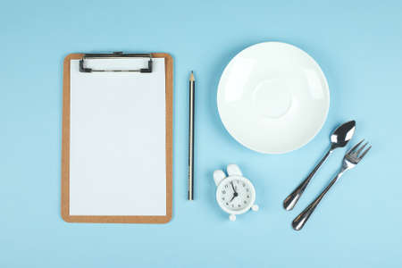 Intermittent fasting, ketogenic, diet concept. Empty plate with white alarm clock and white note notepad on blue background. Flat lay, top view, mockupの写真素材