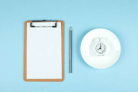 Intermittent fasting, ketogenic, diet concept. Empty plate with white alarm clock and white note notepad on blue background. Flat lay, top view, mockupの写真素材