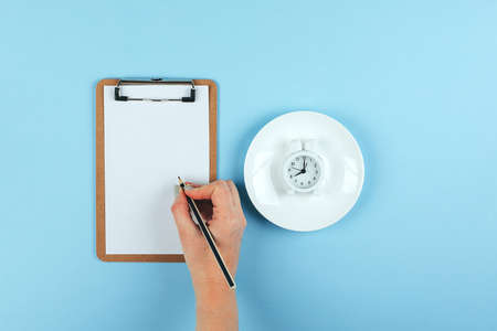 Intermittent fasting, ketogenic, diet concept. Female hand writting weight loss planÑ Empty plate with white alarm clock on blue background. Flat lay, top viewの写真素材