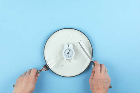 Intermittent fasting, ketogenic, diet concept. Empty plate with white alarm clock on blue background. Flat lay, top viewの写真素材