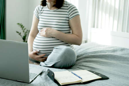 Beautiful pregnant young woman working on a laptop sitting on the bed in the bedroom. Home office, home education. Social distance during quarantine, self-isolation concept.の写真素材