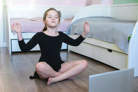 Schoolgirl watching online video on laptop and doing sports exercises - yoga, gymnastics, choreography. Stay at home. Concept of self isolation, quarantine, online education.の写真素材