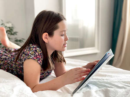 Young school girl reading a book on bed. Home education, stay home. Children leisure in quarantine...