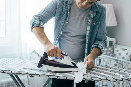 Young father ironing baby clothes in bedroom. Concept of housework, chores at home, domestic life in quarantine period. Stay home.の写真素材