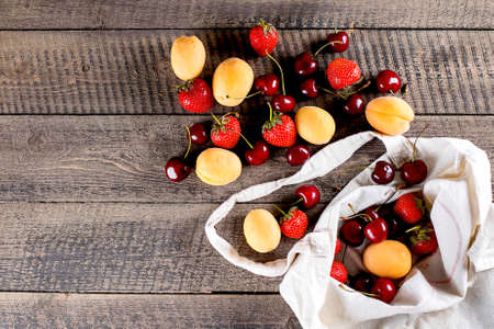 Reusable bag with fruits and berries. Tote bag, minimal waste. Apricot, strawberry and cherry in fabric bag on wooden table background. Top view, copy space.の写真素材