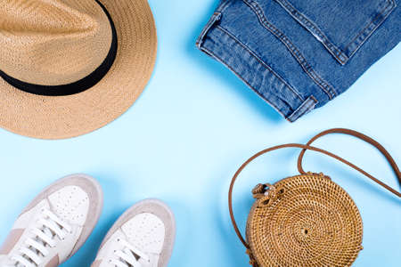 Blue jeans shorts, sneakers, hat and modern rattan bag on blue background. Flat lay, top view, copy...