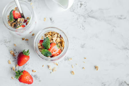 Two jars with tasty parfaits made of granola, strawberries and yogurt on white marble table. Top...