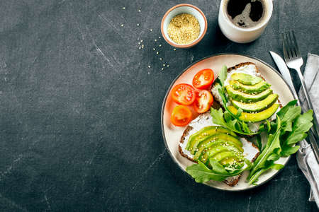 Avocado toasts with cottage cheese, arugula, cup of coffee on dark table. Healthy breakfast, snack....