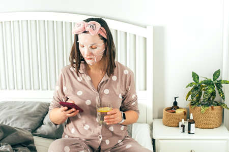 Mature women in sheet mask in pajama spending cozy morning during isolation period, reading book.の写真素材