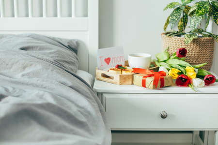 Breakfast for mom on bedside table. Coffee, delicious breakfast, bouguet of flowers tulips, gift box. Home light interior, modern interior, bedroom with gray linens on bed.の写真素材