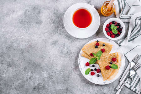 Delicious Crepes Breakfast on gray concrete table background. Orthodox holiday Maslenitsa. Pancakes with berry black currant, raspberry, jar of honey and mint. White cup of tea. Top view, copy spaceの写真素材