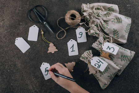 Preparation of christmas advent calendar. Female hand writing number on label note. Presents wrapped in fabric eco bags. Flat lay, top viewの写真素材
