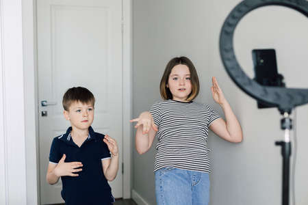 Happy teen girl and boy 7-8 years dancing at camera filming video using smartphone on tripod LED lamp at home. Concept of young blogger.の写真素材