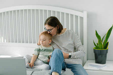 Young single mother mom with toddler boy son sitting on bed using laptop, searching web, watching cartoons together. Concept of work from home, freelancerの写真素材