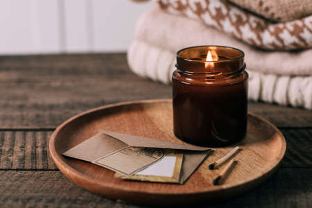 Burning candle in small amber glass jar with wooden wick, stack knitted season sweaters. Cozy lifestyle, hygge concept. copy spaceの写真素材