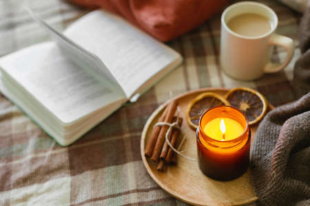Burning candle with wooden wich in amber glass gar, open book and cup of coffee or tea. autumn home decor. Cozy fall mood.の写真素材