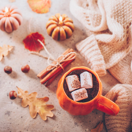 Autumn comfort beverage. Mug with cacao with marshmallow on table, cozy warm sweater, candle and autumn leaves.の写真素材