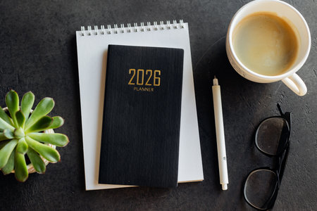 A tidy workspace featuring a 2026 planner, spiral notebook, white pencil, glasses, and a cup of...