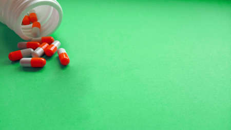 Red-white pharmaceutical medicine pills, capsules spread out of white plastic drug bottle on green...