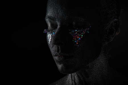 Conceptual art Portrait of Beautiful Woman in Black makeup with Sparkles on a Face isolated on a dark backgroundの写真素材