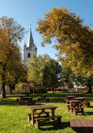 Targu Mures, Romania - October 17. 2025. Picnic area with wooden tables in the green fortress park, framed by autumn trees with yellow leaves and the historic Reformed Church steeple in the backgroundのeditorial素材