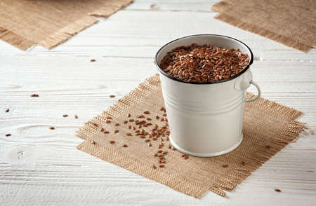 flax seeds in a small enameled bucket on a tissue napkin on a white wooden background