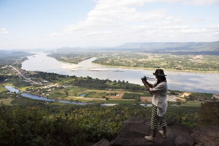 Thai woman travel visit and posing take photo on ridge stone of cliffs at Wat Pha Tak Suea temple with view of landscape of Nongkhai city and loas and Mekong river in Nong Khai, Thailandの写真素材