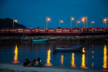 Thai women and friend sit relax playing mobile smart phone on sand beach with people playing skateboard Saphan saranwithi red bridge in night time on September 6, 2021 in Prachuap Khiri Khan, Thailandのeditorial素材