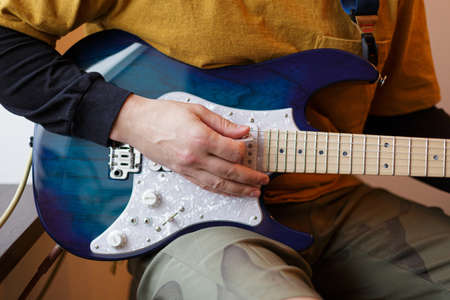 A young guy plays an electric guitar sitting by the window