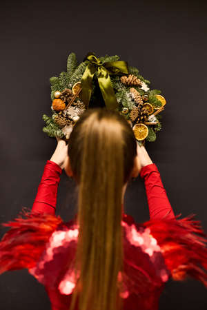 A girl in an elegant red dress hangs a Christmas wreath on the wallの写真素材