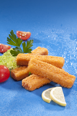 Breaded fried Fish fingers with lettuce and cherry red tomato with lemon slice on blue background...