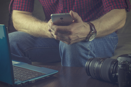 Hipster in a red shirt and blue jeans sits with a smartphone on the couch. Next to a young man notebook and SLR camera.の写真素材
