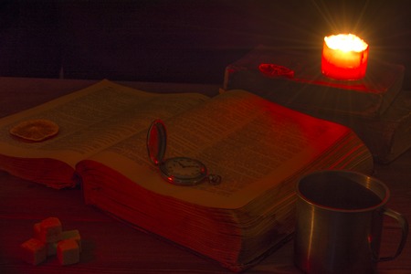 Old book on the table. The open Holy Bible is on the table. Vintage pocket watch. A burning candle over the Bible illuminates its pages. Mug, pieces of sugar and orange slices.の写真素材