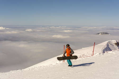 Rosa Khutor, Russia March 20, 2020. A snowboarder walks along the track on rose Peak above the cloudsのeditorial素材