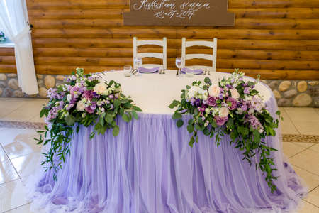 a beautifully designed festive wedding table with a composition of white roses, greenery and purple flowers, and in the background-a wooden wall with a garland of light bulbs.Purple color schemeの写真素材