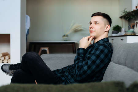 Handsome happy young man smiles and holds his hand to his chin, guy in green shirt in a cage, at home, conceived to implement the idea.gray sofa in the interior, against the background of the kitchenの写真素材