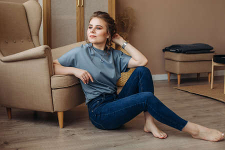 A young girl sits on the floor, leaning on an armchair, at home, barefoot, looking to the side. In blue clothes, a beige armchair, a light and stylish interior.の写真素材