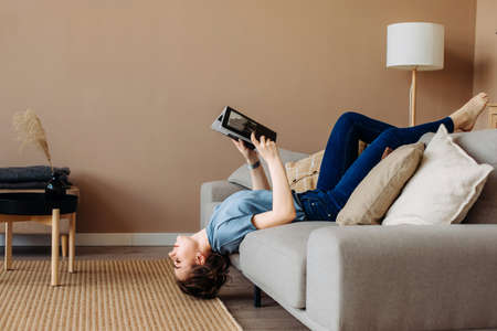 Girl reading a book lying on the couch upside down in the living room at home, romantic mood. Calm stylish interior. The woman in blue casual clothes. Lifestyleの写真素材