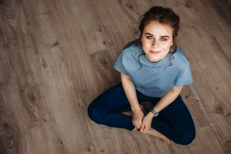 A young girl is sitting on a wooden floor, barefoot, looking at the camera. In a blue blouse and jeans. Beautiful makeup, collected hairstyle. view from above. Copy spaseの写真素材