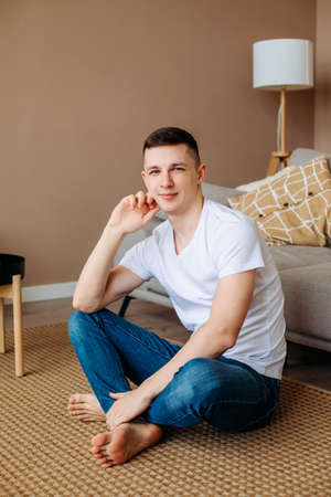 A young guy at home sitting on the floor in a beige interior barefoot and looking at the camera....